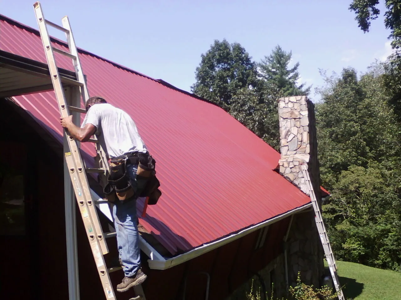 Roofer on a ladder beside a red metal roof and stone chimney — Soffit & Fascia Repair by Roofing Brothers in Kincaid